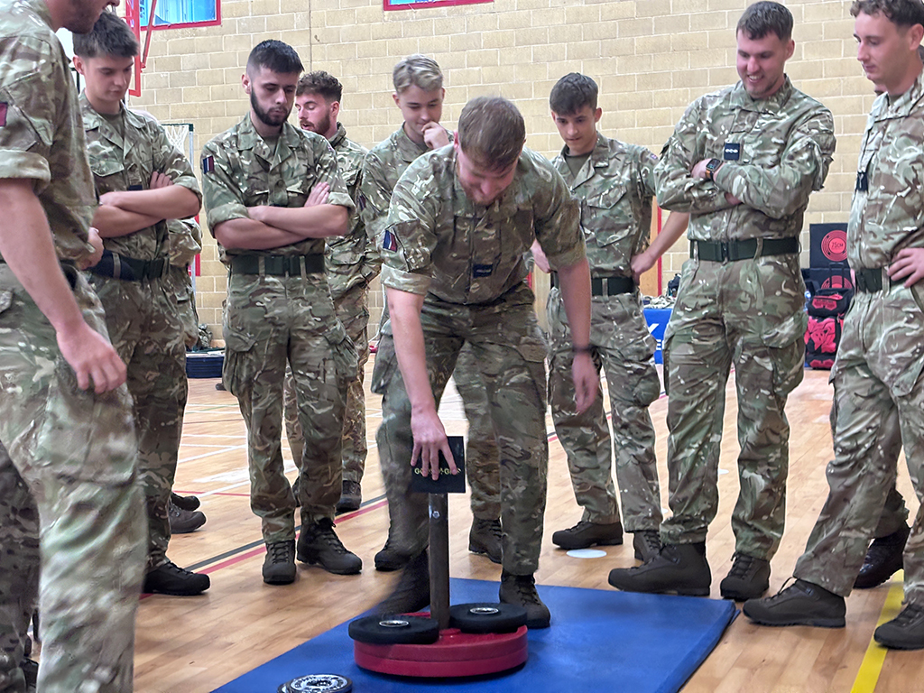 Military personnel watch a demonstration of a weight lifting challenge.